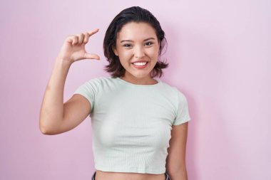 İspanyol kökenli genç bir kadın pembe arka planda durup gülümsüyor ve el hareketleriyle el hareketleriyle küçük boy işareti yapıyor ve kameraya bakıyor. ölçüm kavramı. 