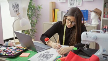 Odaklanmış İspanyol bir kadın, iyi donanımlı bir terzi dükkanında, dikiş malzemeleri ve renkli ipliklerle bir tasarım çiziyor..