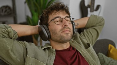Rahat, genç, İspanyol bir adam rahat bir oturma odasında kulaklıklarla müzik dinliyor..