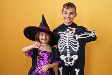 İyimser erkek ve kız kardeşler ürkütücü cadılar bayramı kostümleri giyiyorlar, heyecan ve neşeyle işaret ediyorlar, sarı izole edilmiş bir zemin üzerinde neşeli gülücükler saçıyorlar.