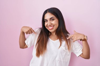 Pembe arka planda duran genç Arap kadın yüzünde bir gülümsemeyle gururla ve mutlu bir şekilde kendini işaret ediyor.. 