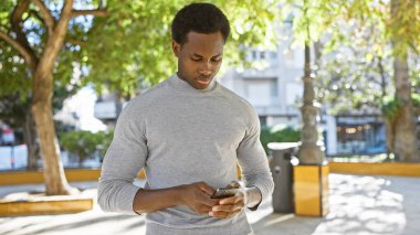 Ağaçlarla çevrili güneşli bir şehir parkında akıllı telefonuyla mesaj atan Afrikalı genç bir Amerikalı..