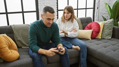 Bir kadın, rahat bir oturma odasında video oyunu kontrolörüne odaklanmış bir adama endişeli bakar..