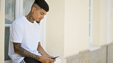 Havalı latin adam akıllı telefona dalmış, güneşli şehir arka planında ciddi bir ifadeyle yazı yazıyor.