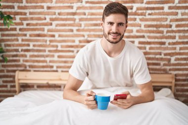 Beyaz bir adam akıllı telefon kullanıyor. Yatak odasında kahve içiyor.