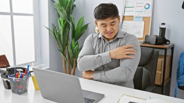Modern ofis ortamında dizüstü bilgisayarı üzerinde çalışırken omuz ağrısı çeken Asyalı genç bir adam..