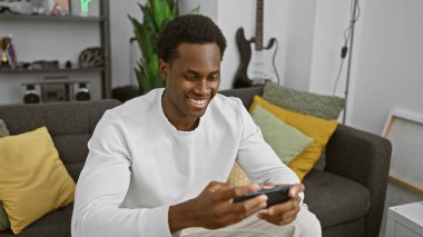 Modern bir oturma odasında otururken akıllı telefon kullanan gülümseyen Afrikalı genç bir Amerikalı..