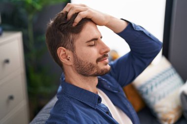 Kafkas 'lı genç adam saçını tarıyor, elleriyle evdeki kanepede oturuyor.