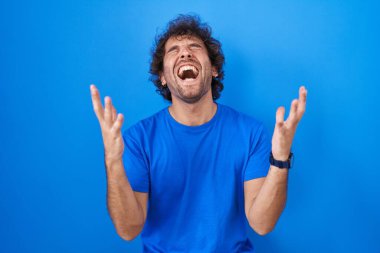Hispanic young man standing over blue background celebrating mad and crazy for success with arms raised and closed eyes screaming excited. winner concept 