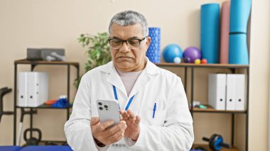 Beyaz laboratuvar önlüklü olgun bir adam rehabilitasyon merkezinin içinde akıllı telefon kullanıyor ve arka planda egzersiz ekipmanları var..