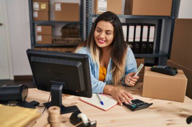 Young beautiful plus size woman ecommerce business worker counting dollars using calculator at office