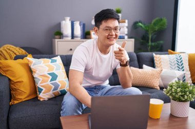 Genç Asyalı adam evinde dizüstü bilgisayar kullanıyor kanepeye oturmuş mutlu ve komik yüzüyle kamerayı işaret ediyor. iyi enerji ve titreşimler. 