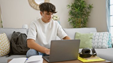 Konsantre olmuş genç İspanyol erkek öğrenci, çevrimiçi eğitime dalmış, rahat oturma odası kanepesinde oturuyor, dizüstü bilgisayara odaklanmış, evinde dinleniyor, teknolojiyle çevrili.