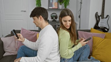 Bir kanepede sırt sırta verip, rahat bir oturma odasında akıllı telefonlar kullanan erkek ve kadın..