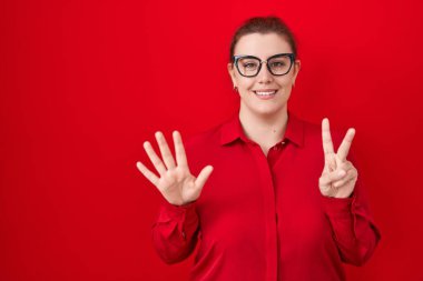 Kırmızı arka planda dikilen kızıl saçlı genç İspanyol kadın yedi numaralı parmaklarıyla gülümseyerek kendinden emin ve mutlu bir şekilde onu işaret ediyor.. 