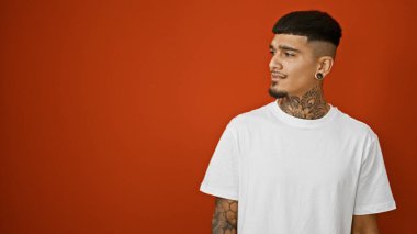 Havalı ve kararlı, genç dövmeli Latin adam ciddi bir şekilde aşağı bakarken rahat bir poz veriyor, ateşli kırmızı bir arka planda izole edilmiş, sıradan ama yoğun bir yüz ifadesi takınıyor..