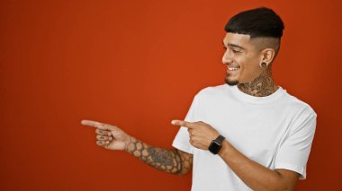 Neşeli genç latin adam kendinden emin, kendine güvenen, enfeksiyon derecesinde mutlu bir gülümseme sergilerken dövmeli ellerini göstererek, canlı, izole bir kırmızı arkaplanı işaret ediyor..