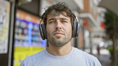 Havalı sarışın sakallı genç adam şehir ritminde kaybolmuş, dışarıda duruyor, müziğe dalmış kulaklıklarından üflüyor, ciddi şekilde odaklanmış görünüyor, güneşli şehir manzarasında banyo yapıyor..