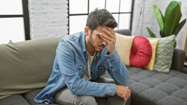 Modern bir dairede koltukta oturan sakallı, kederli, genç bir İspanyol adam, elleriyle yüzünü kapatıyor..