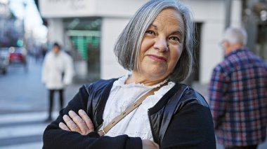 Gri saçlı, kollarını kavuşturmuş, hareketli bir caddede kameraya bakan, kendinden emin bir kadın..