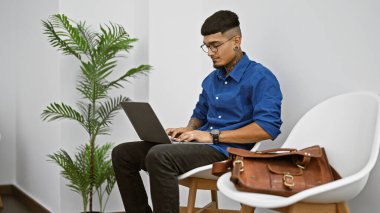 Ciddi genç Latin bir adam, dövmeli bir profesyonel. Modern işçilerin mücadelesinin bir sembolü olan bekleme odasında otururken dizüstü bilgisayarına hiç şüphesiz konsantre oluyor..