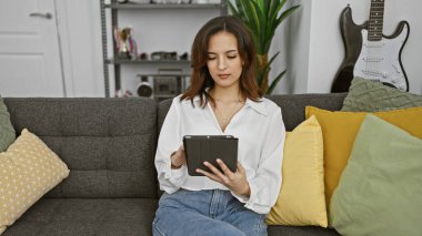 Şık bir iç mekan giymiş genç bir İspanyol kadın kanepenin üzerinde tabletine konsantre oluyor..