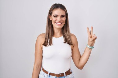 Beyaz arka planda duran Latin kökenli genç bir kadın iki numarayı gösterip işaret ederken kendinden emin ve mutlu bir şekilde gülümsüyor.. 