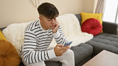 Yakışıklı, ispanyol bir adam üzgün bir şekilde kanepede oturuyor. Kendini evinde rahat bir şekilde otururken akıllı telefon kullanmaya kaptırmış.