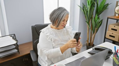 Olgun bir kadın modern bir ofiste akıllı telefonunu karıştırıyor. Kapalı alan bitkileri ve çağdaş dekorasyonları var..