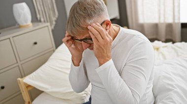 Gri saçlı, gözlüklü, stresli bir adam yatakta oturuyor..