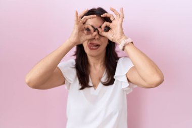 Orta yaşlı İspanyol kadın, günlük beyaz tişört ve gözlük takıyor dürbün gibi jest yapıyor, dili dışarı çıkarıyor, gözleri parmaklarından bakıyor. çılgın ifade. 