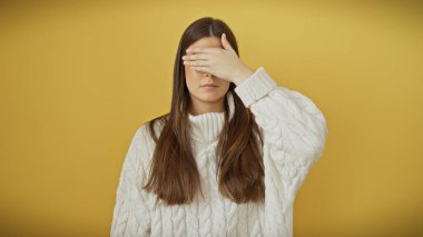 Genç bir kadın, elini sarı arka planda tutarak gözlerini kapatır, mahremiyet ya da şaşkınlık hissi uyandırır..