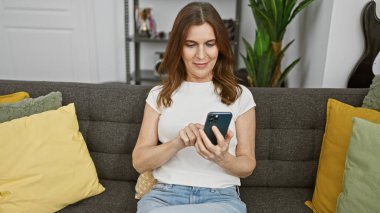 Orta yaşlı bir kadın rahat oturma odasındaki koltukta akıllı telefon kullanıyor.