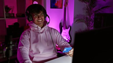 Çekici, genç İspanyol bir adam, neşeyle gece boyu oyun oynuyor! Geniş bir gülümseme, elinde kartla, süslenmiş oyun odasında dijital alışverişe hazır.