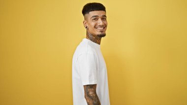 Bulaşıcı bir gülümsemesi ve havalı dövmesi olan kendinden emin genç Latin bir adam, gelişigüzel ayakta duruyor, izole edilmiş sarı bir arka plana neşe ve pozitiflik saçıyor..