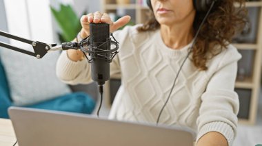A middle-aged woman adjusts a microphone in a radio studio while working on a podcast. clipart