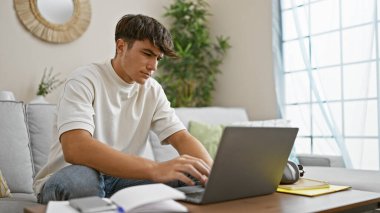 Konsantre olmuş genç İspanyol erkek öğrenci, çevrimiçi eğitime dalmış, rahat oturma odası kanepesinde oturuyor, dizüstü bilgisayara odaklanmış, evinde dinleniyor, teknolojiyle çevrili.