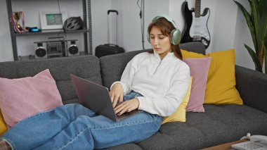 Genç bir kadın rahat bir oturma odasında gri bir kanepede uzanırken dizüstü bilgisayar kullanıyor..