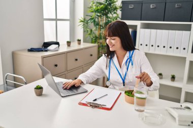 Genç ve güzel İspanyol doktor klinikte dizüstü bilgisayar kullanarak salata yiyor.