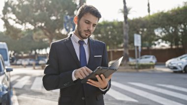 Şehir caddesinde ağaçlar ve arabalarla tablet kullanan yakışıklı İspanyol takım elbiseli adam.