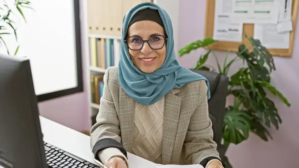 戴着眼镜和头巾在现代化的办公室里工作的中年妇女笑着 表现出专业精神和自信 — 图库照片