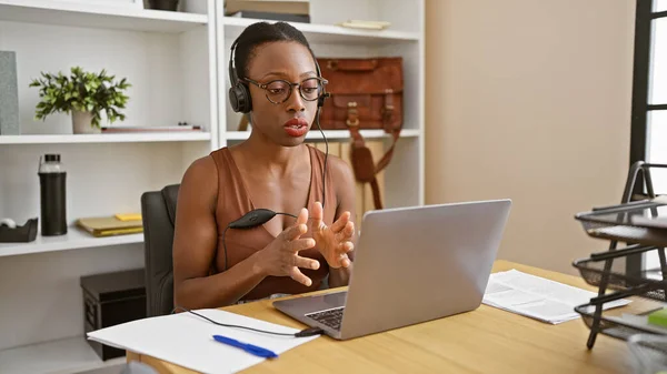 Beautiful african american woman boss wearing glasses, heartily engaged in an online business call. working indoors in her office, she\'s relaxed yet serious, managing her duties via her laptop.