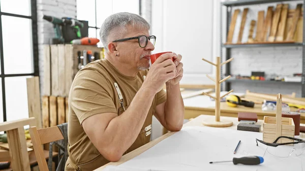 Middle-aged man drinks coffee in a carpentry workshop, reflecting on woodwork with tools around.