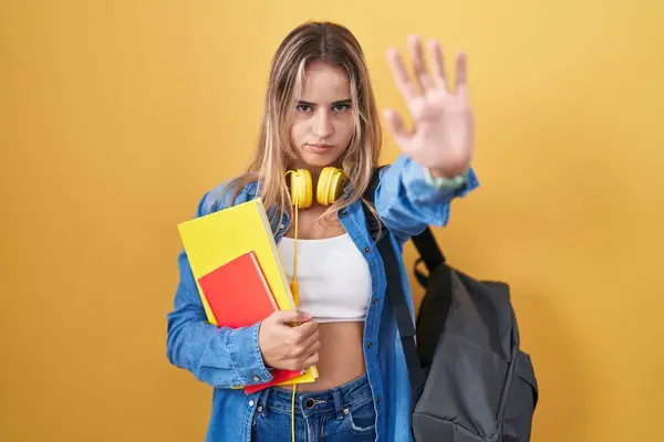 Young blonde woman wearing student backpack and holding books doing stop sing with palm of the hand. warning expression with negative and serious gesture on the face.