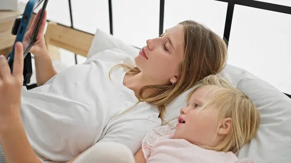 Caucasian mother and daughter lying on bed watching video on touchpad at bedroom