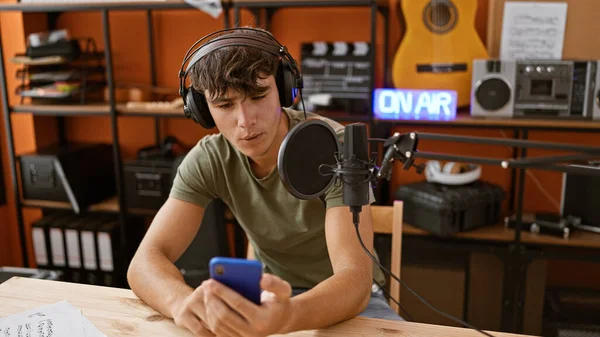 Young hispanic teenager, charismatic radio reporter, masterfully reading news information live on air from smartphone in bustling radio studio