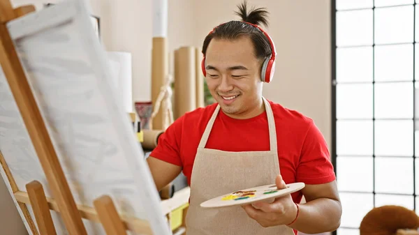 Smiling, young chinese man artist, a handsome adult with a pigtail hairstyle and apron, creatively drawing on a canvas in art studio while intimately listening to his favorite song on headphones.