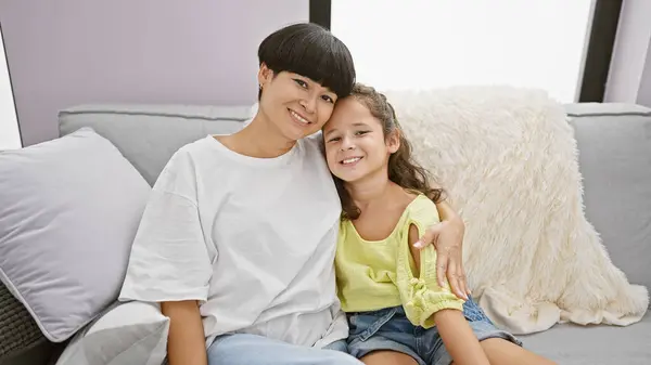 Confident mother and daughter sharing a joyful hug, smiling heartily while sitting on the living room sofa at home  a beautiful expression of family love