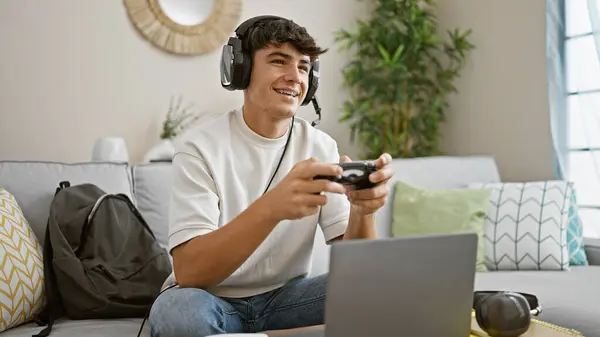 Cheerful, young hispanic teenager student, gamepad in hand, engrossed in online gaming, lounging on living room sofa, making his apartment a lively gaming house.