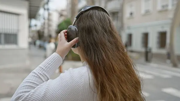 カジュアルな服装の若いヒスパニック女性が街路に立っている間にヘッドフォンを調整 — ストック写真